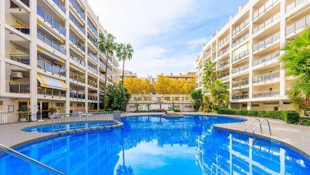 A swimming pool surrounded by multi-story residential buildings with balconies, palm trees, and a clear sky.