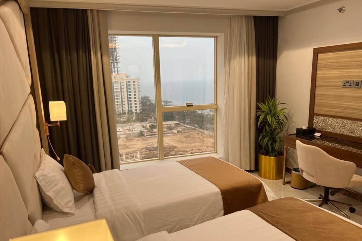 A hotel room with two beds, a desk, a chair, a large window with a view of buildings, and a potted plant.