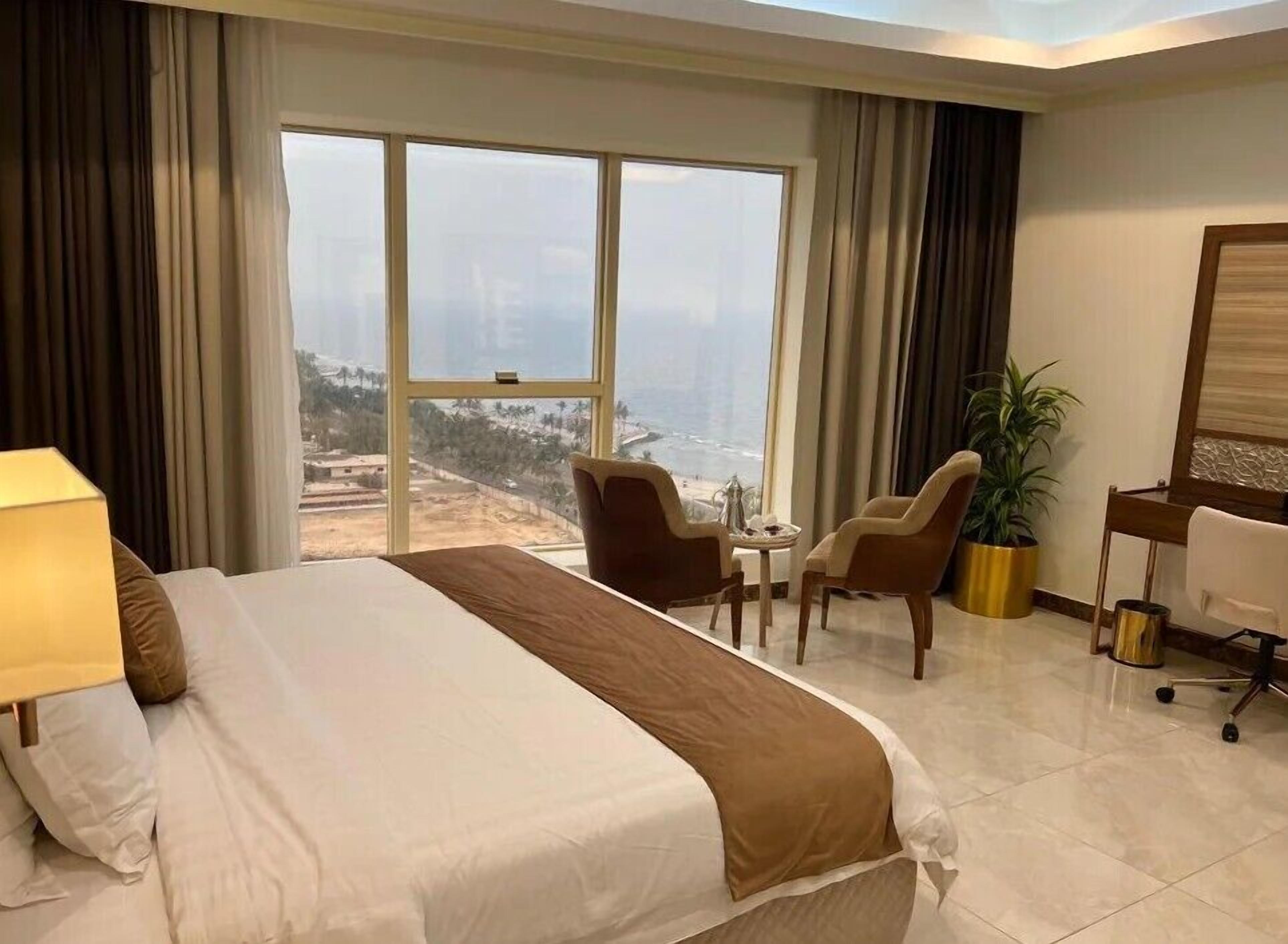 A hotel room with a large bed, a desk, two chairs, a potted plant, and a view of the ocean.