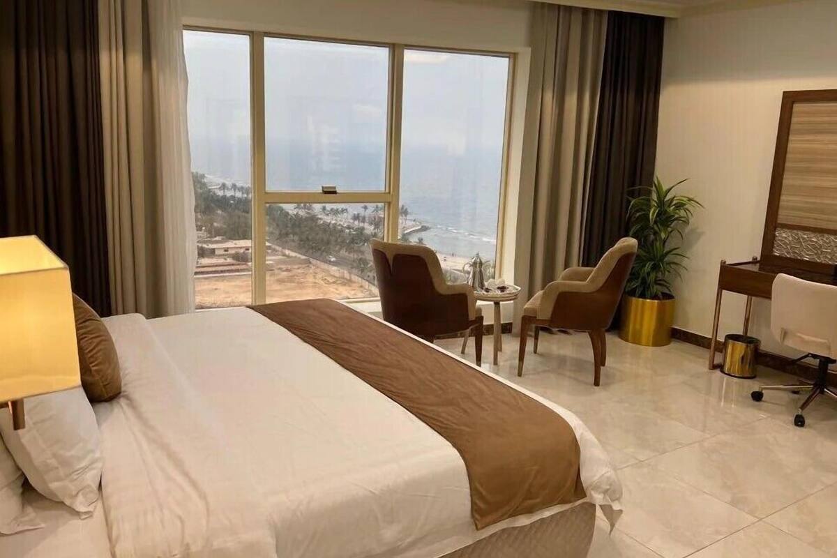 A hotel room with a large bed, a desk, two chairs, a potted plant, and a view of the ocean.