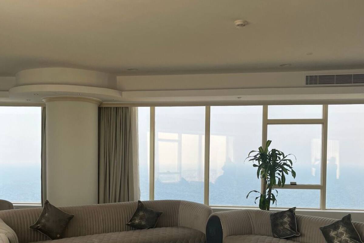A spacious living room with a large window offering a view of the sea and a bridge. There is a beige sofa with decorative pillows and a potted plant.