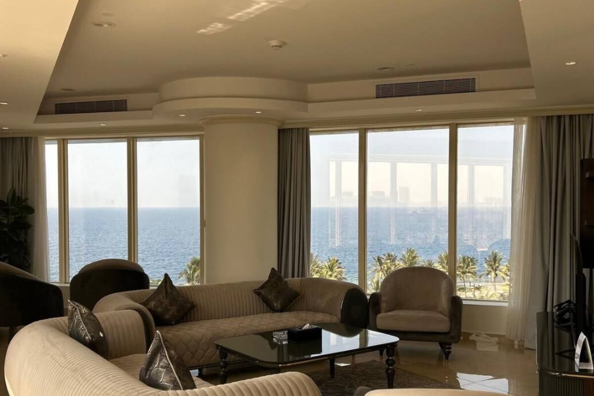 A spacious living room with a large sofa, armchairs, and a coffee table. The room has large windows offering a view of the ocean and a city skyline.