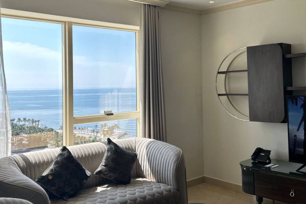 A modern room with a large window offering a view of the ocean and palm trees. There is a comfortable sofa with cushions and a small desk with a telephone and a television.