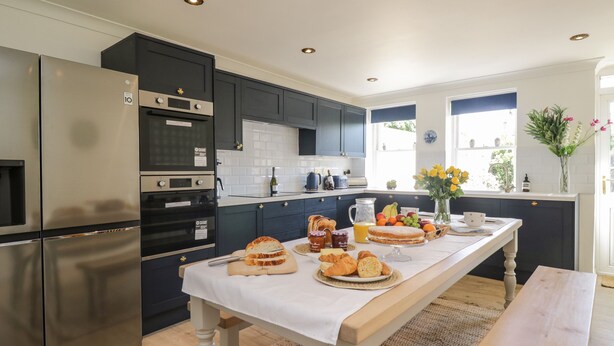 A modern kitchen with stainless steel appliances, a wooden table set with breakfast items, and a view of the outdoors through large windows. - The Admiral's Home (Whitehaven)