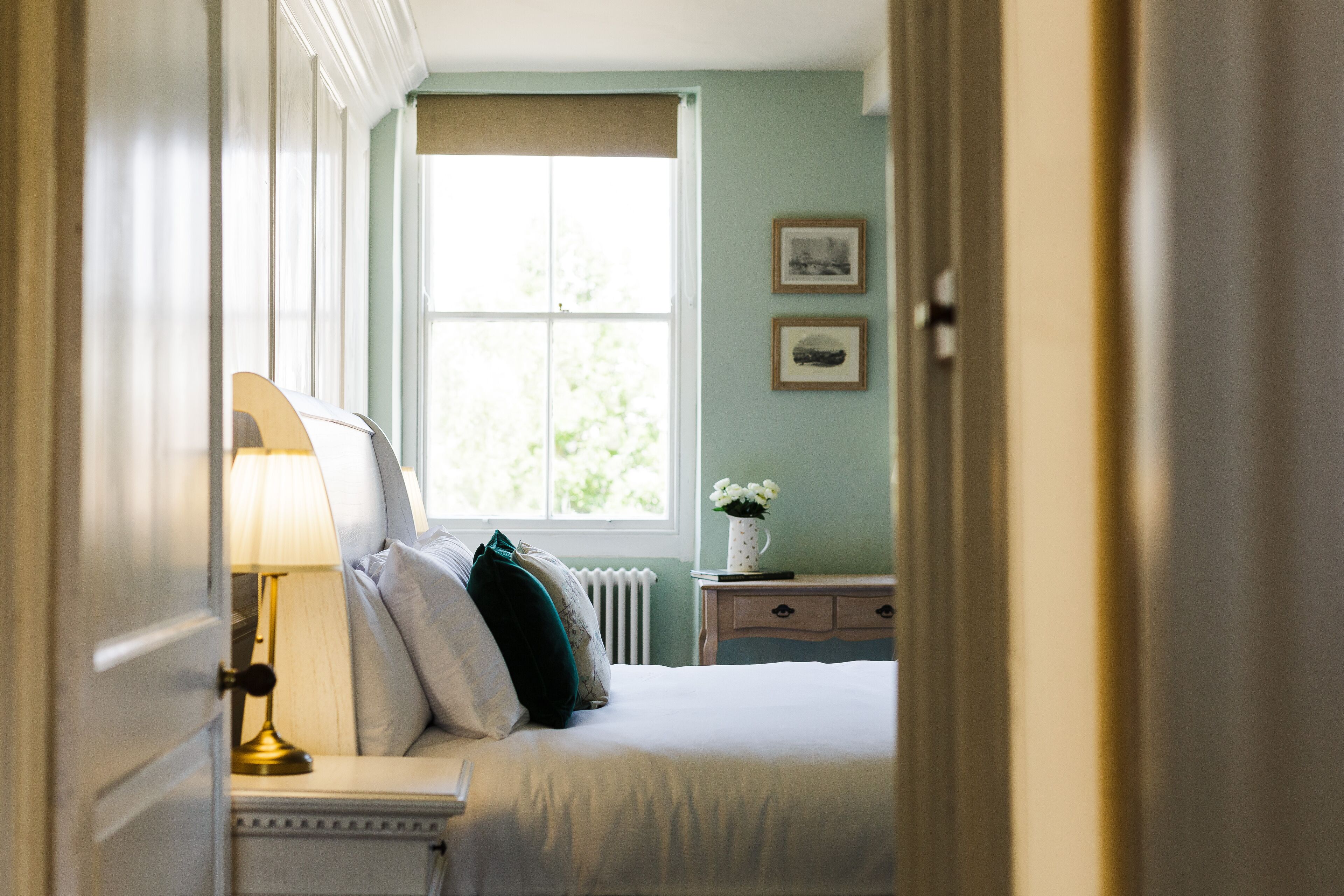 A bedroom with a bed, bedside table, lamp, and window with a view of greenery.