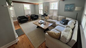 Un salón con un sofá color beige, una mesa de centro de madera y un sillón de cuero negro. Hay ventanas con contraventanas blancas, un cuadro en la pared y una lámpara sobre una mesita auxiliar.