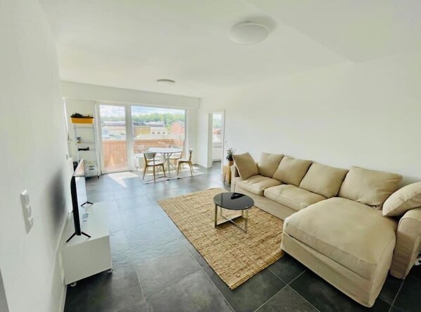 A modern living room with a beige sofa, a small round table, and a large window offering a view of the city. - Large 1 bedroom with Terrace & Parking (Luxembourg City)