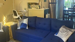 A modern living room with a blue sectional sofa, a small dining table, and a kitchen area with a refrigerator and microwave.