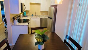 A compact kitchen with white cabinets, a stainless steel refrigerator, and a dining table with a potted plant.