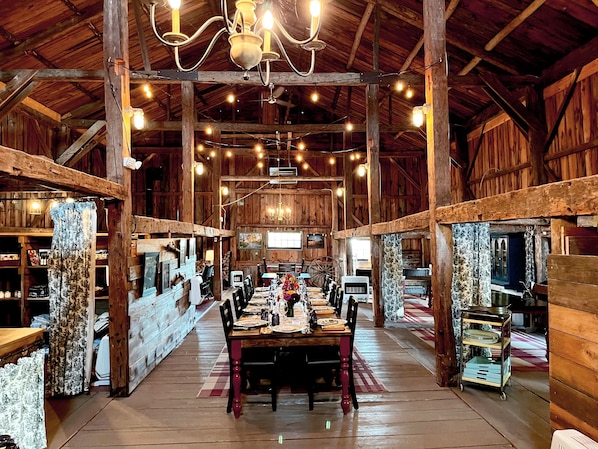 A rustic barn-style dining area with a long wooden table set for a meal, surrounded by wooden walls and beams, and a chandelier hanging from the ceiling.