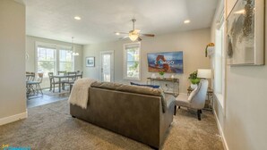 A spacious living room with a large gray sofa, a dining area with a table and chairs, a ceiling fan, and a TV mounted on the wall. - Mountain View Daybreak Home | Pets Welcome (South Jordan)