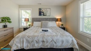 A bedroom with a large bed, two bedside tables with lamps, a dresser, and a window with blinds. - Mountain View Daybreak Home | Pets Welcome (South Jordan)