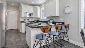 A modern kitchen with white cabinets, a stainless steel oven, and a central island with bar stools. - Mountain View Daybreak Home | Pets Welcome (South Jordan)