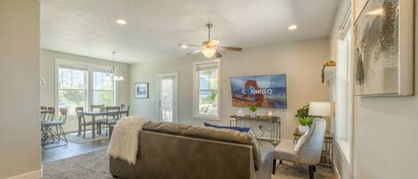 A spacious living room with a large gray sofa, a dining area with a table and chairs, a ceiling fan, and a TV mounted on the wall.