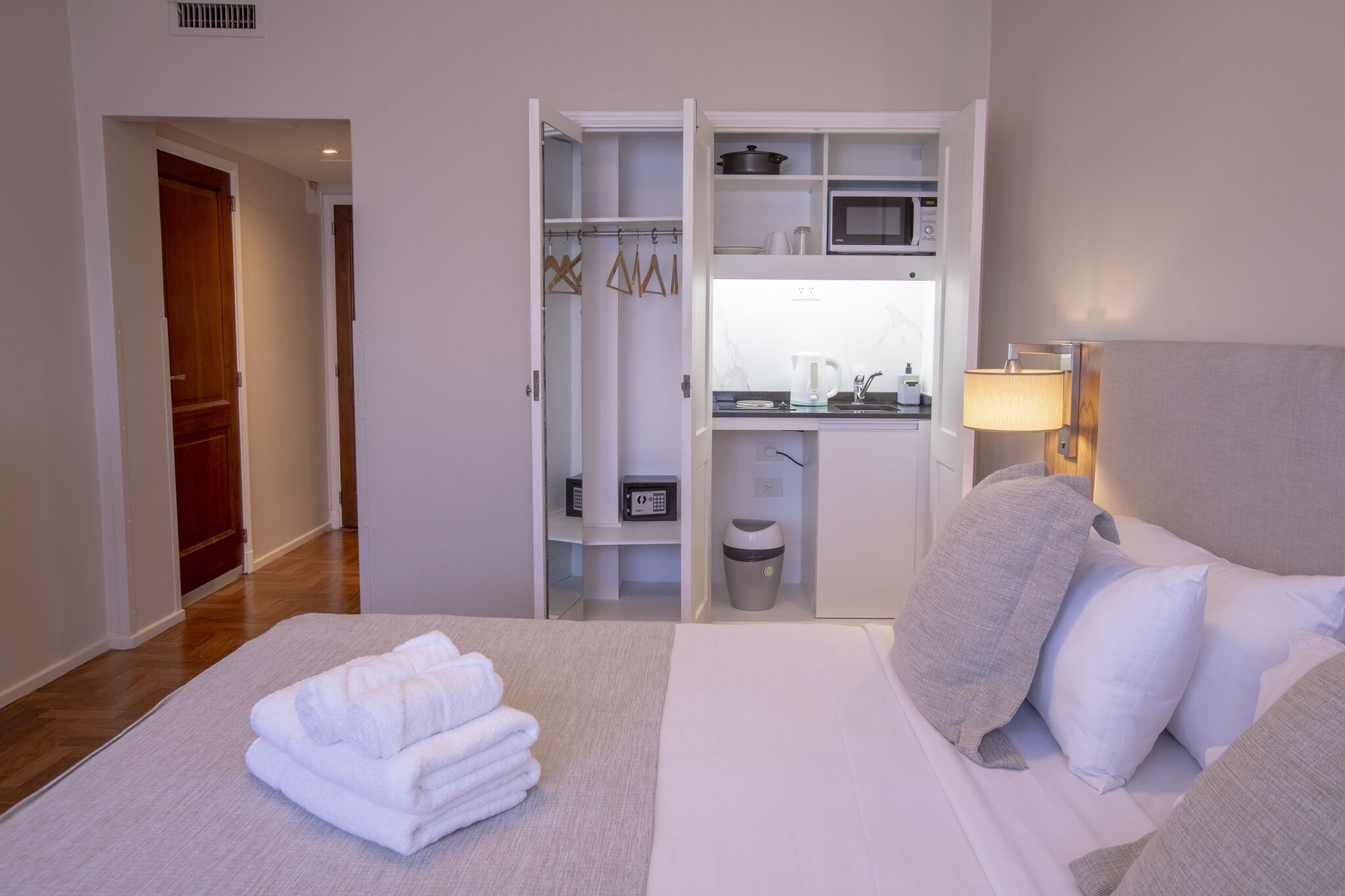Quarto de hotel com cama arrumada, closet visível com roupas penduradas, micro-ondas e pequena área de cozinha.