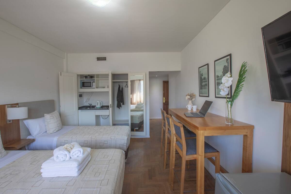 A hotel room with two beds, a kitchenette, a dining area with a table and chairs, and a TV on the wall.