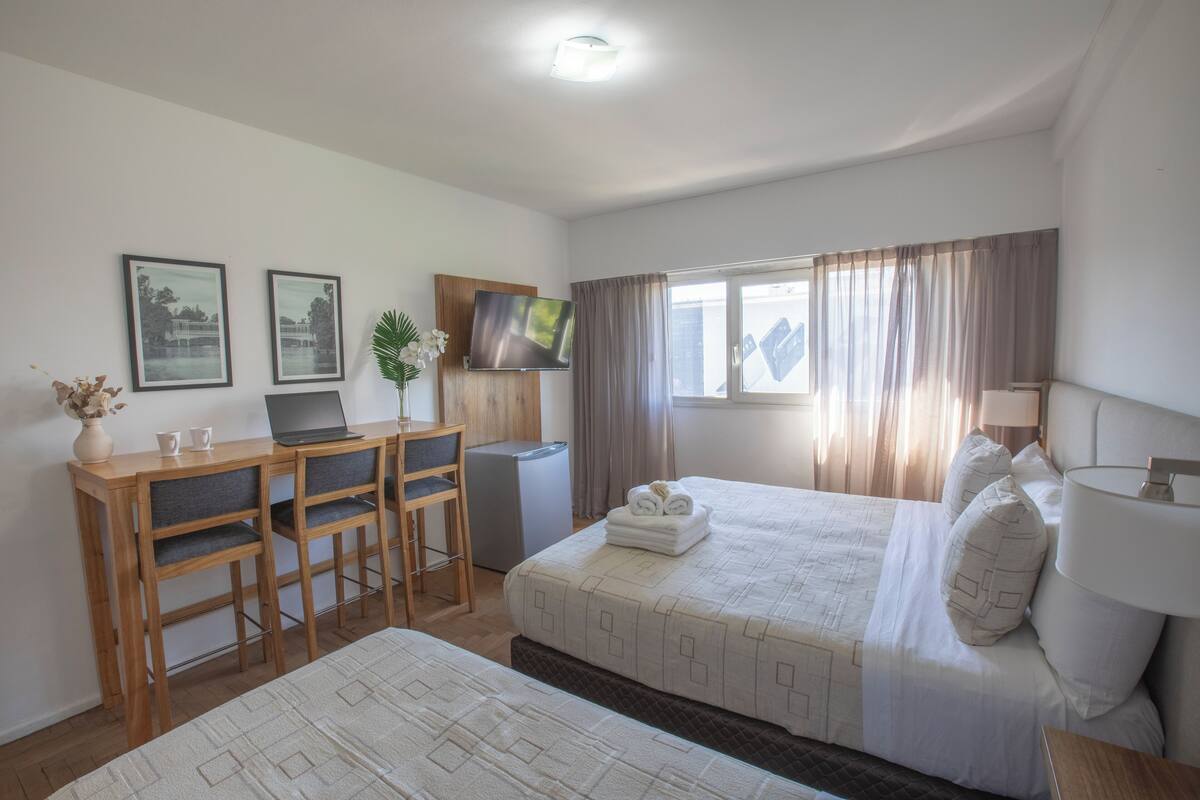 A hotel room with a bed, a desk with two chairs, a TV, and a window with curtains.