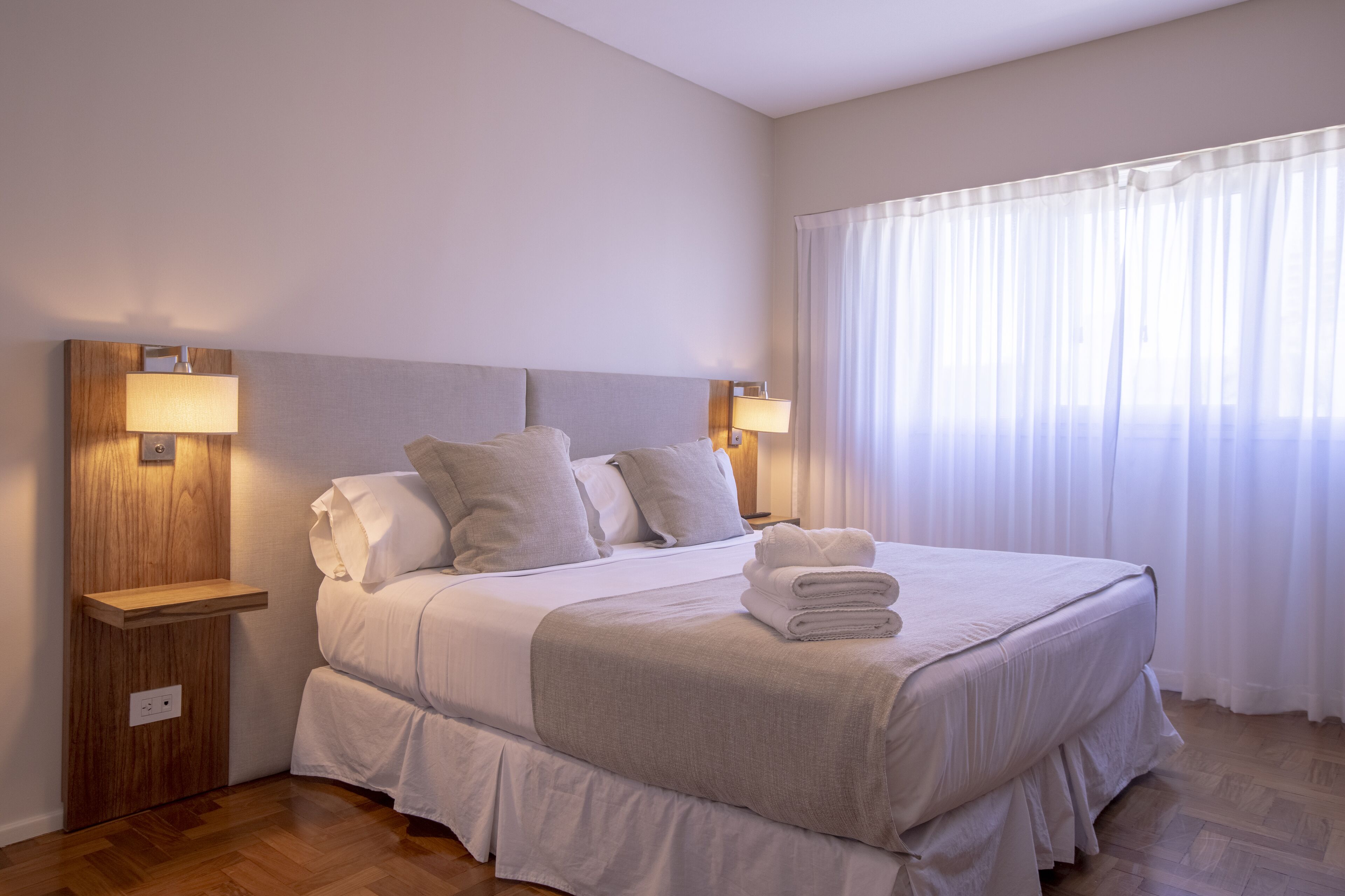 Una cama bien hecha con almohadas y una toalla doblada, una mesita de noche con una lámpara, y una ventana con cortinas de encaje.