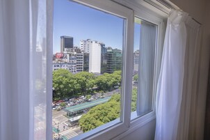 Un paisaje urbano visto a través de una ventana con cortinas blancas, que muestra edificios altos, un parque verde y una calle con vehículos.