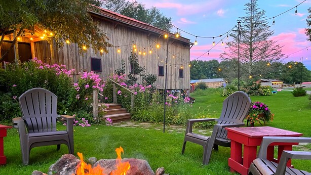 Un'ambientazione rustica all'aperto con un edificio in legno, due poltrone Adirondack, un braciere e luci decorative.