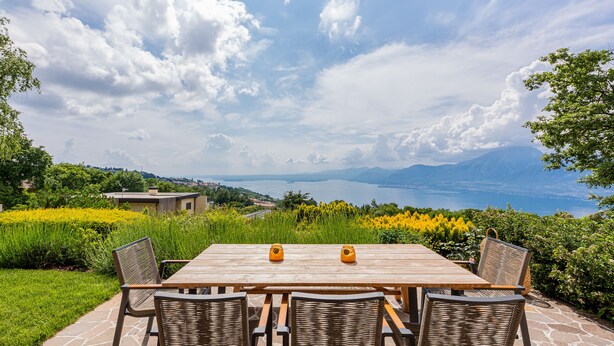 A wooden table with four chairs, set on a stone patio with a view of a lake and mountains.