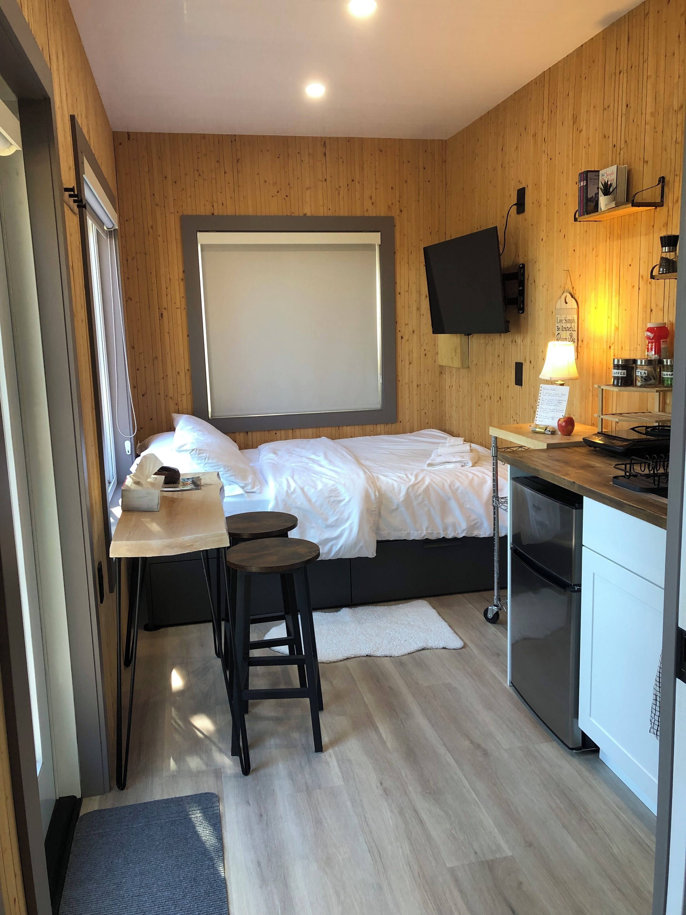 A compact room with a bed, a small desk, a chair, a TV mounted on the wall, and a kitchenette area.