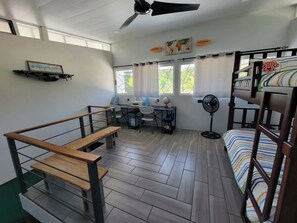 Ein Schlafsaalzimmer mit Etagenbetten, einem Schreibtisch, einem Stuhl, einem Ventilator und einem Fenster mit Vorhängen.