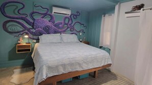A bedroom with a bed, a wall-mounted air conditioner, a nightstand with a lamp, and a wooden floor.