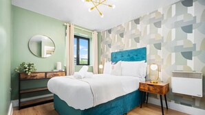A bedroom with a teal bed, a wooden nightstand, a round mirror, and a window with curtains. - 2 Bed Apartment - City Centre - Parking - Balcony
 (Birmingham)