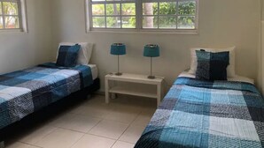A room with two single beds, a small white nightstand with two lamps, and a window with a view of greenery.