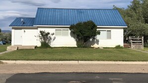 Una casa de un piso con techo de metal azul, revestimiento blanco y un pequeño jardín delantero con un árbol y arbustos.
