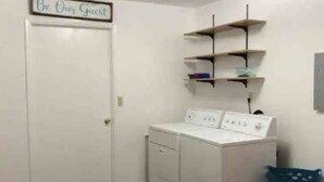 Un cuarto de lavado con lavadora y secadora, una cesta de ropa azul y repisas con objetos encima.