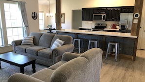 Uma sala de estar moderna com sofá, mesa de centro e área de cozinha com armários escuros, micro-ondas e geladeira.