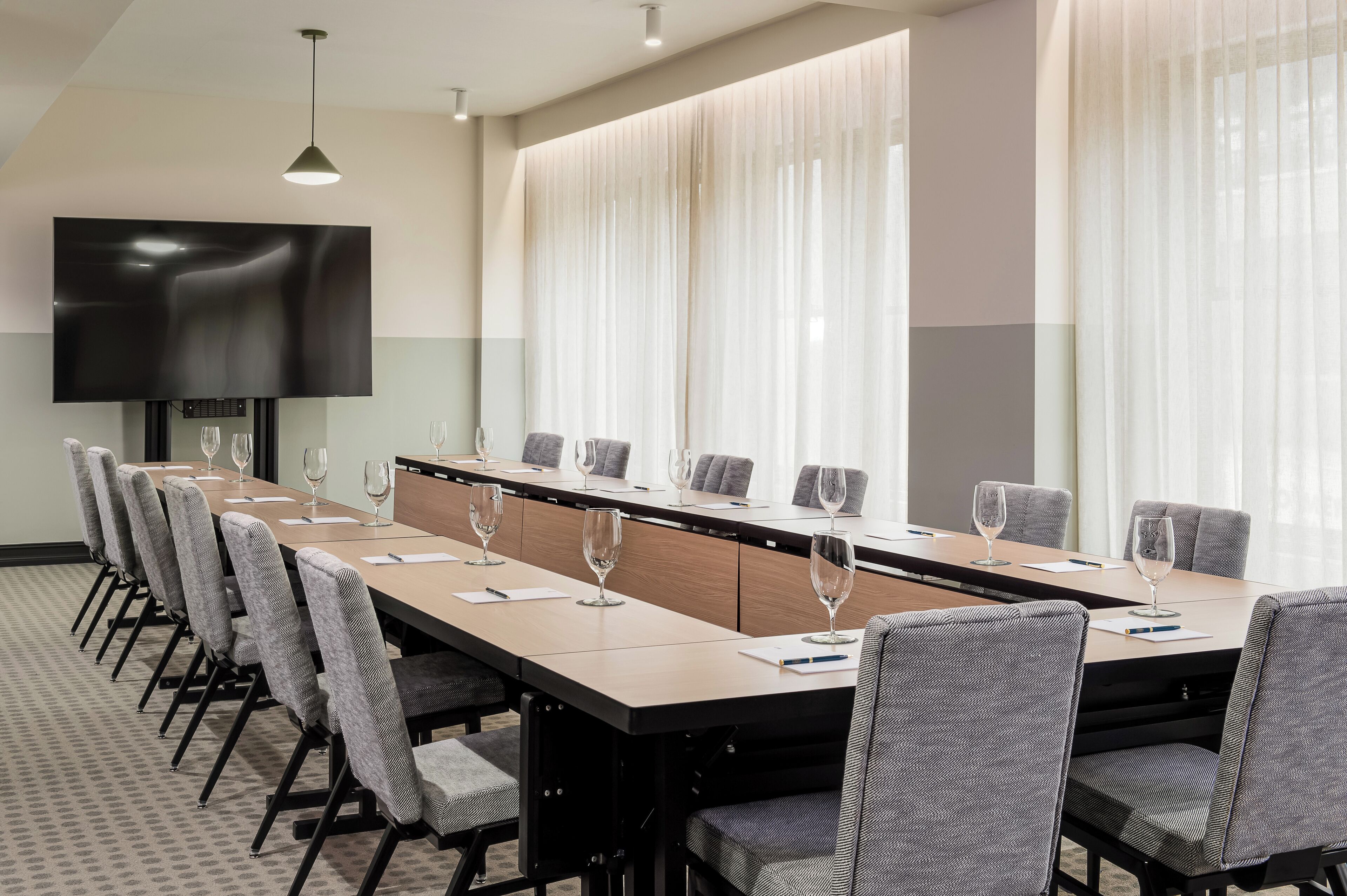 Une salle de conférence dotée d’une longue table, de chaises, d’un téléviseur à écran plat et de grandes fenêtres ornées de rideaux légers.