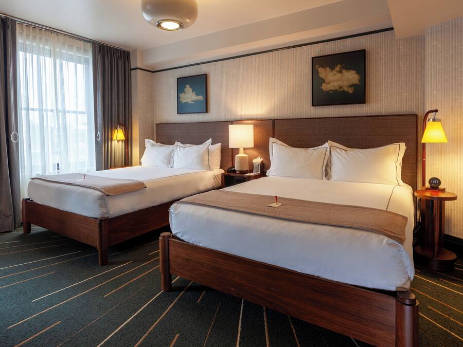 A hotel room with two beds, a large window with curtains, a headboard with artwork, bedside lamps, and a carpeted floor.