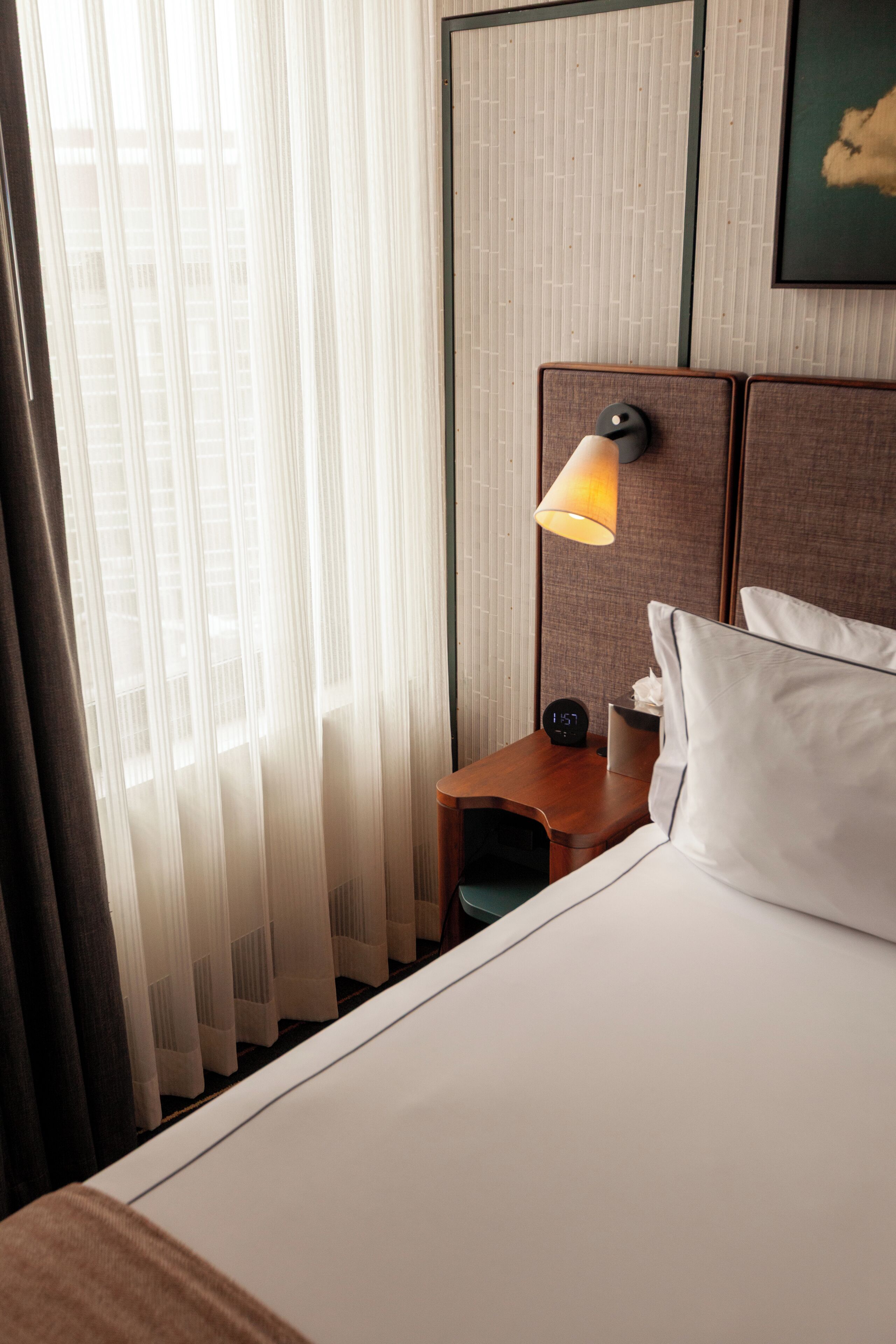 A hotel room with a bed, a nightstand, a lamp, and sheer curtains.