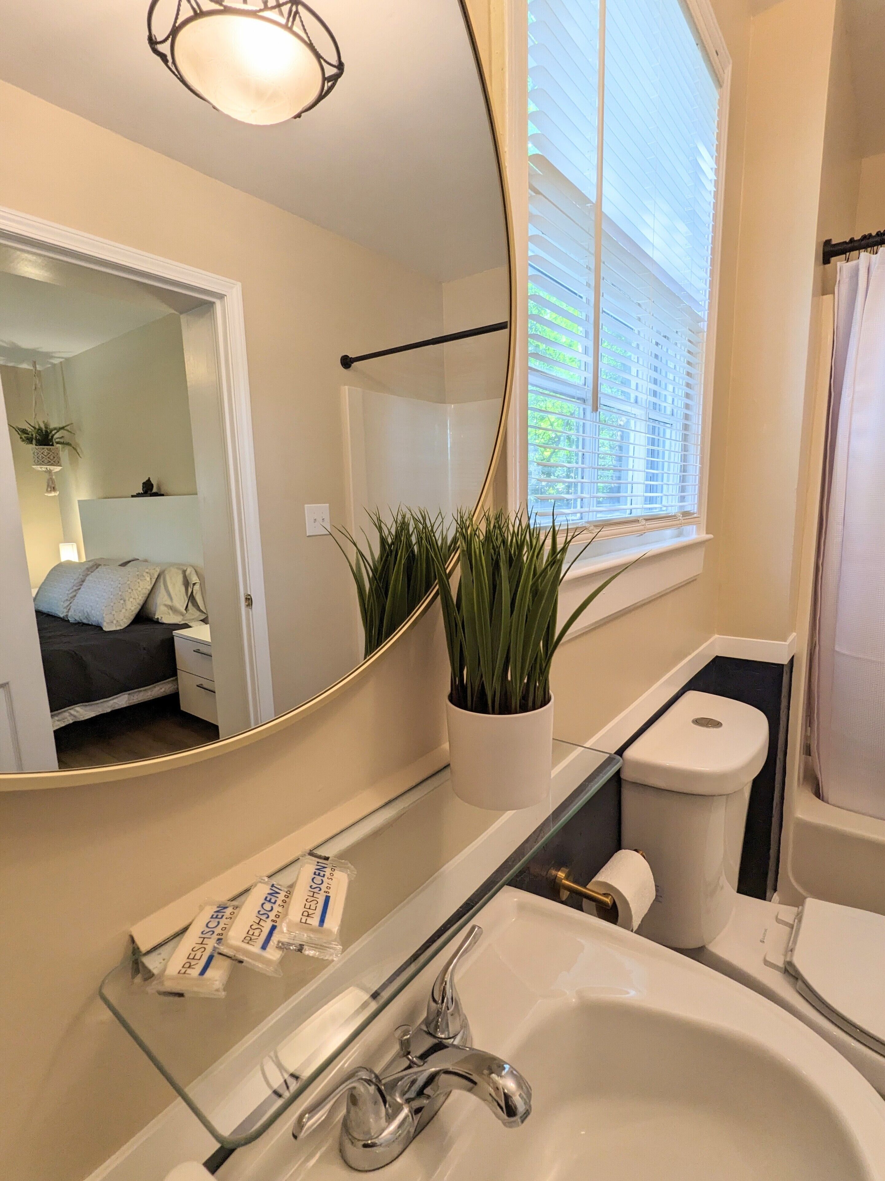 Een badkamer met een wit wastafel, toilet, douche en een grote spiegel. Op het raamkozijn staat een potplant en boven het toilet bevindt zich een handdoekhouder.