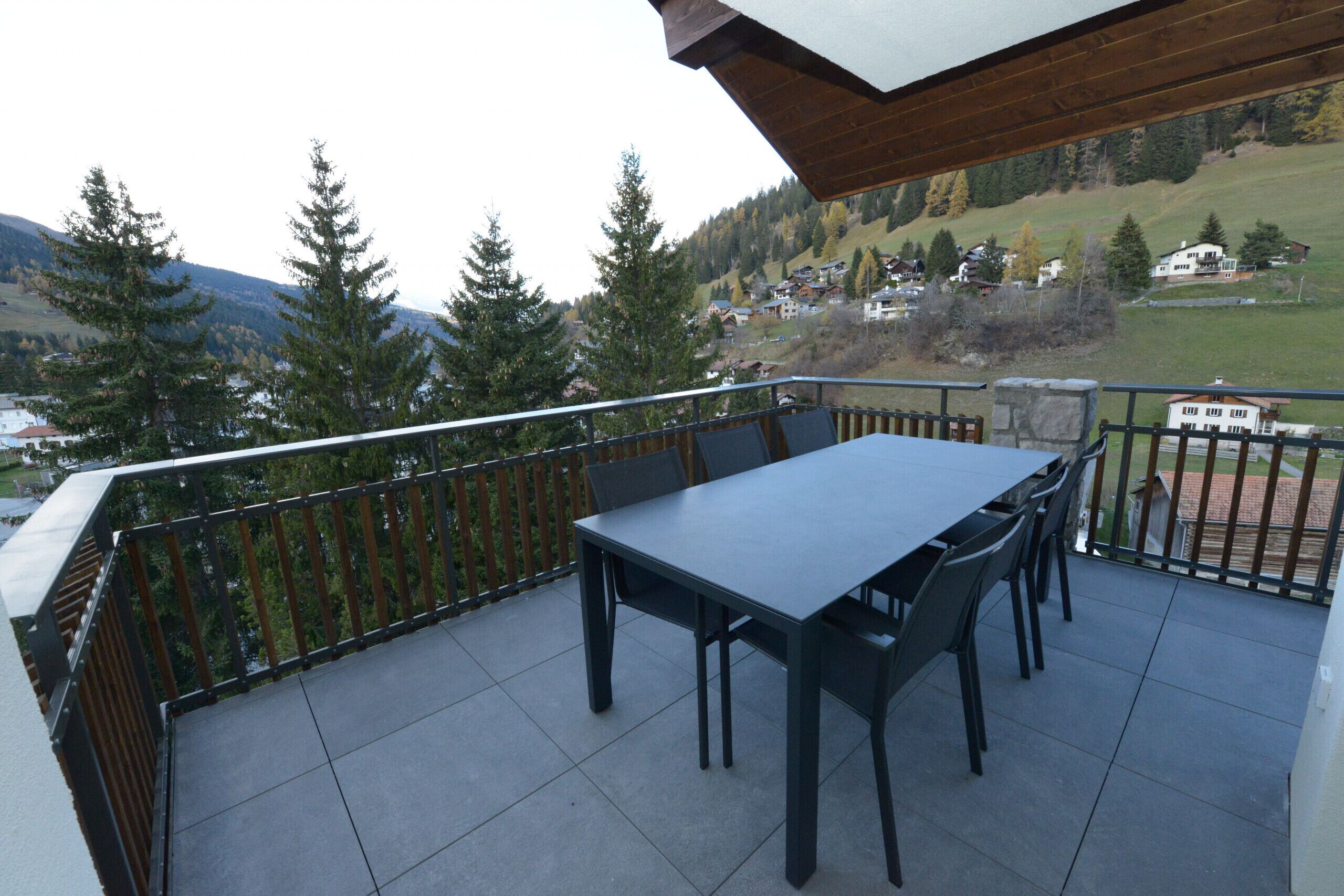 Un balcon avec une table et des chaises, donnant sur un paysage montagneux parsemé de maisons et d’arbres.