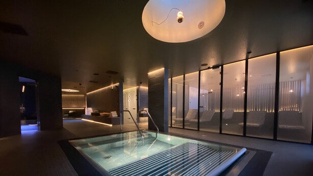 Una piscina interna moderna con parete in vetro, illuminazione a soffitto e area relax con vista sulla piscina.