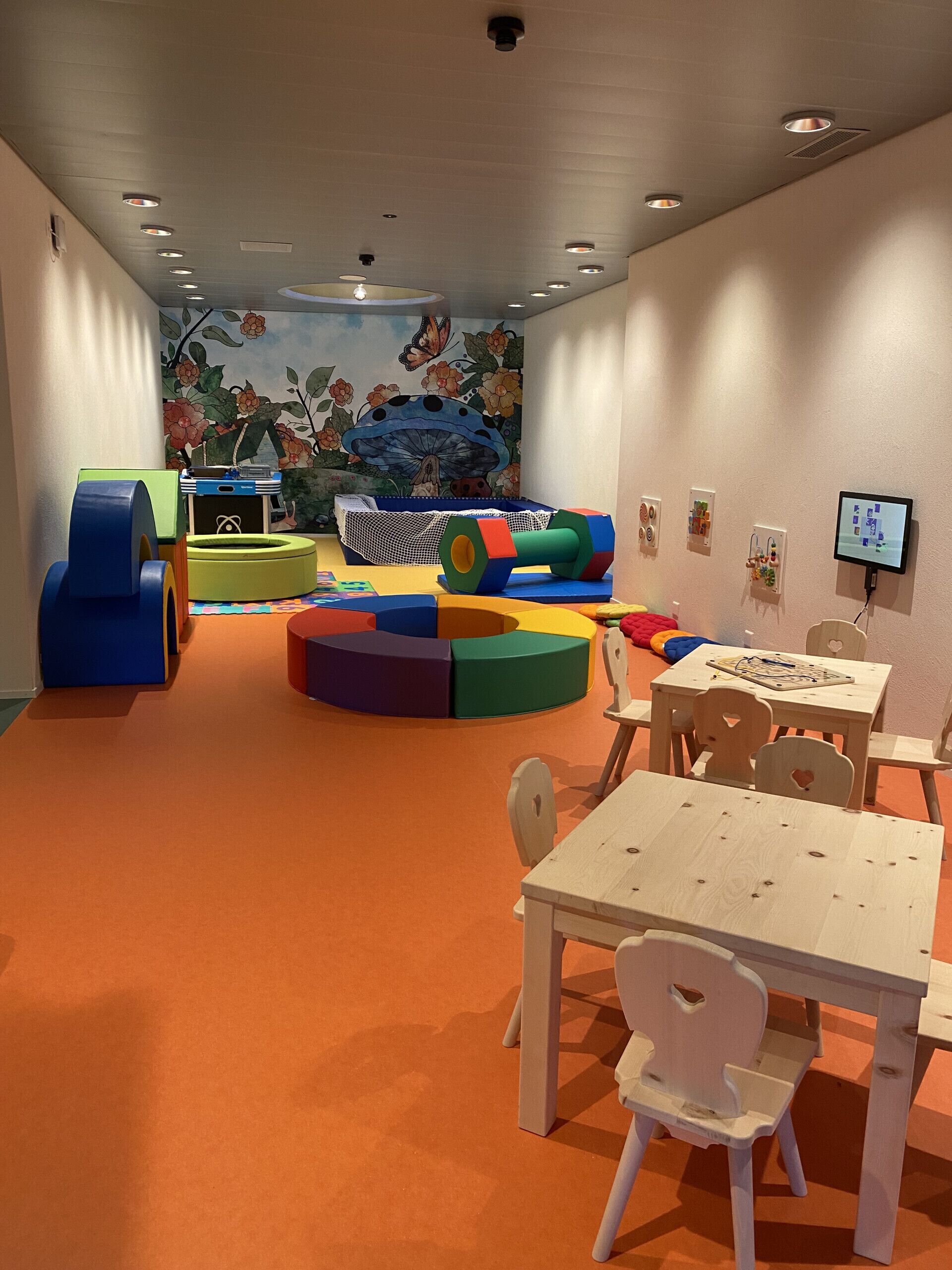 Ein Kinderspielbereich mit bunten Spielgeräten, einer Wandmalerei und einem an der Wand montierten Fernseher.