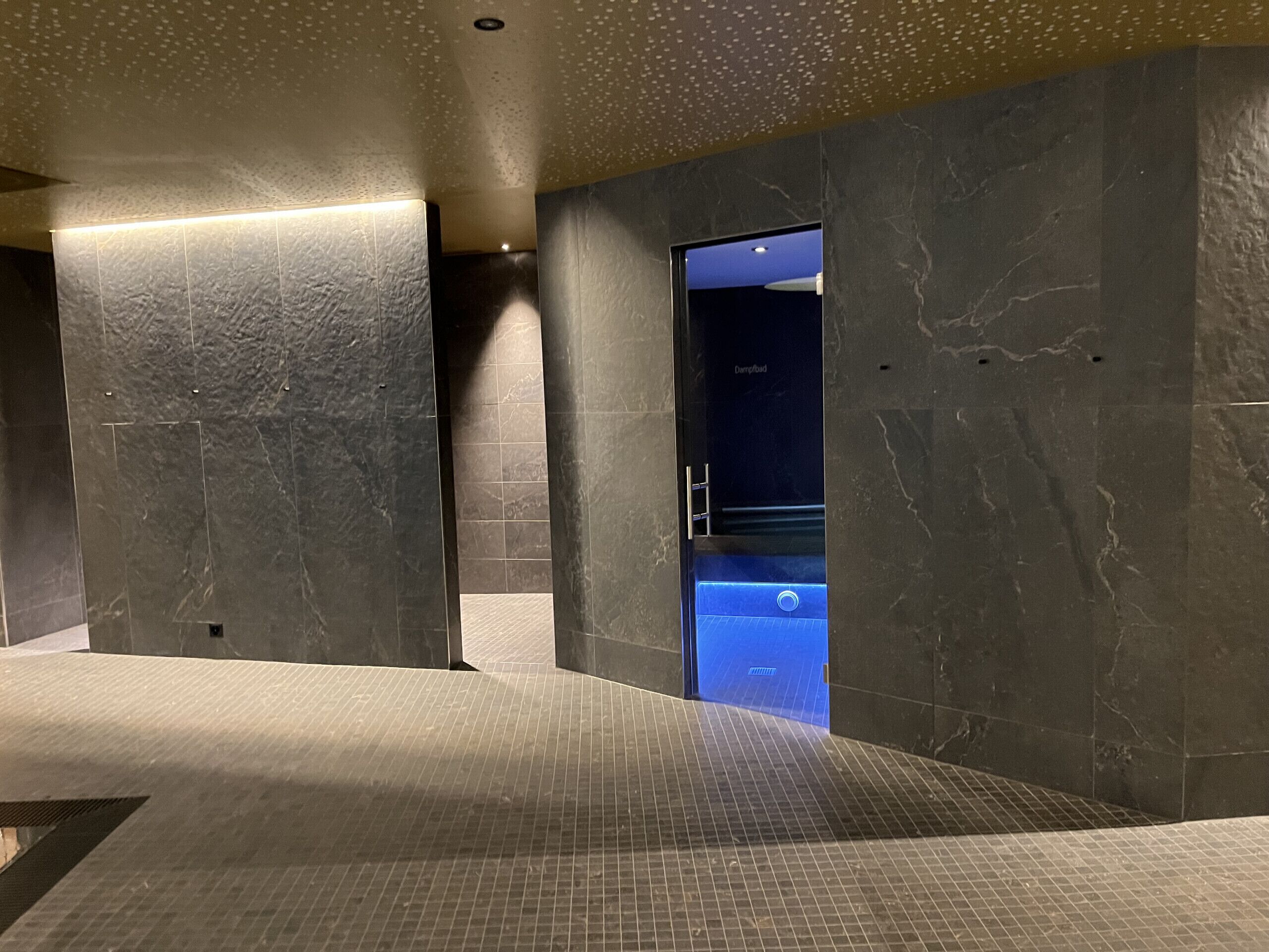 Un'area spa moderna con vasca idromassaggio illuminata di blu, pareti in marmo scuro e pavimento strutturato.