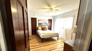 A bedroom with a bed, wooden flooring, a ceiling fan, and a window with curtains.