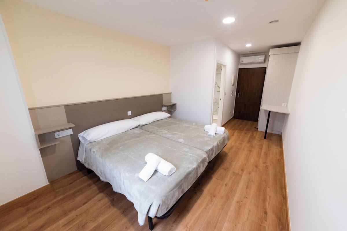 A bedroom with a bed, a nightstand, a wall-mounted shelf, an air conditioning unit, and a door leading to another room.