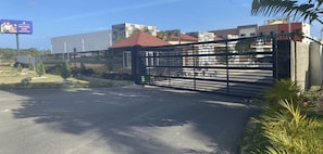 A gated entrance with a metal fence, a red-roofed building, and a billboard in the background. - A cozy and beautiful apartment in Boca Chica (Boca Chica)