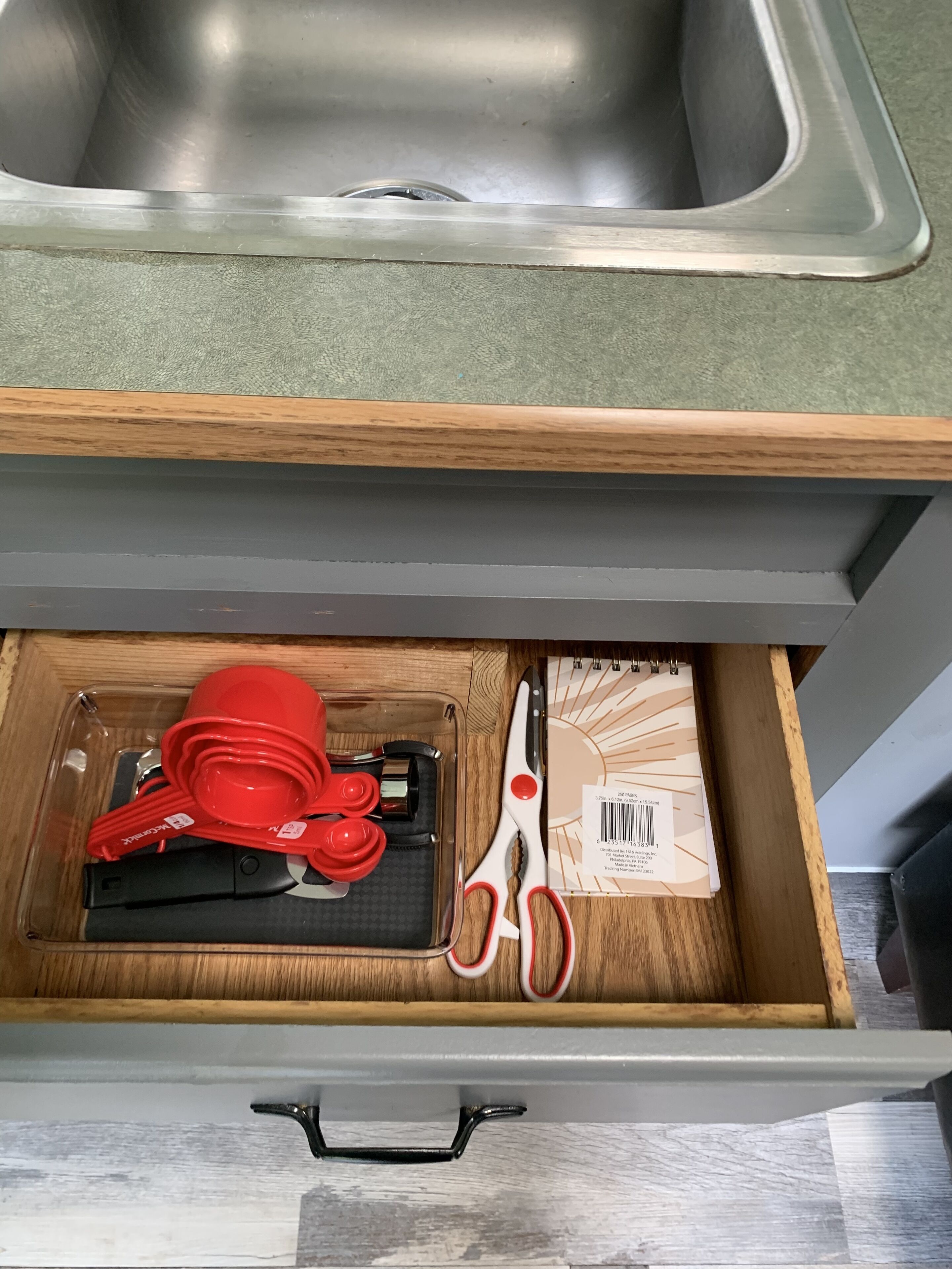 A kitchen with a stainless steel sink and a drawer containing red kitchen utensils, a black appliance, and a spiral-bound notebook.