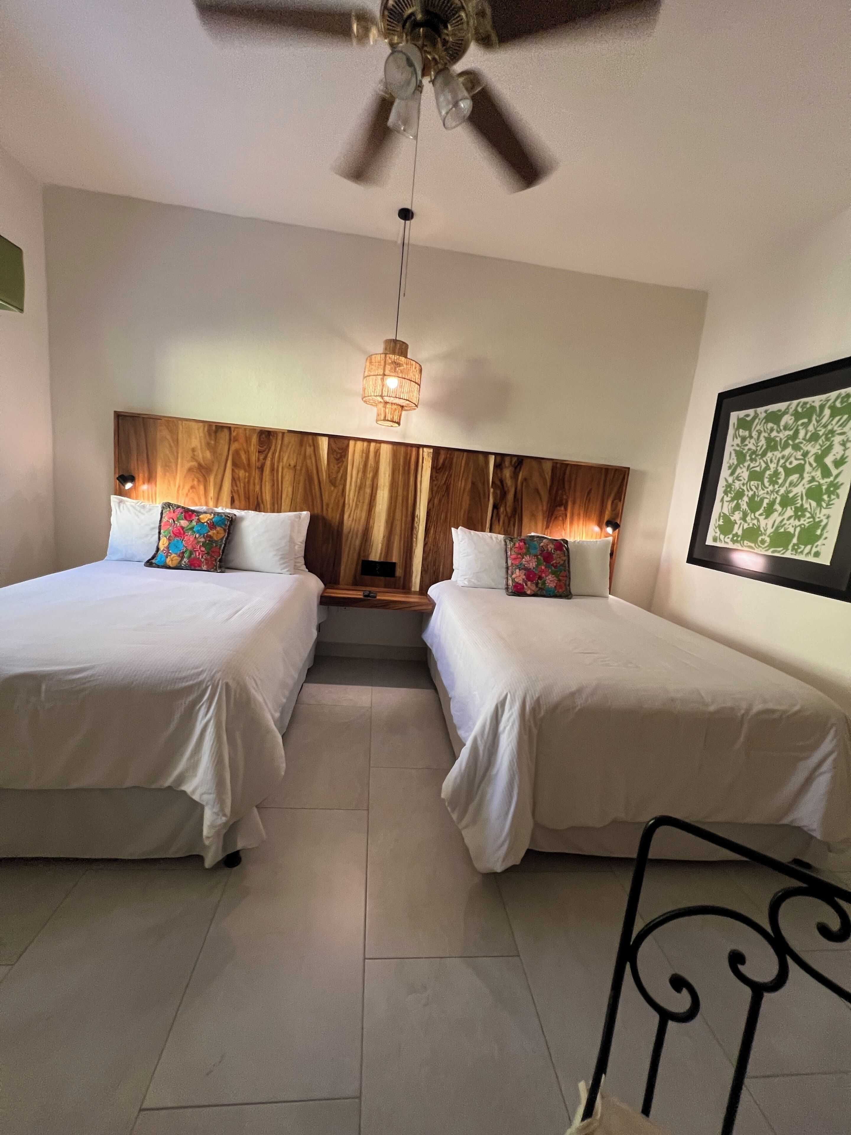 Une chambre d’hôtel avec deux lits, un ventilateur de plafond, une tête de lit en bois, une petite table et une œuvre d’art encadrée au mur.