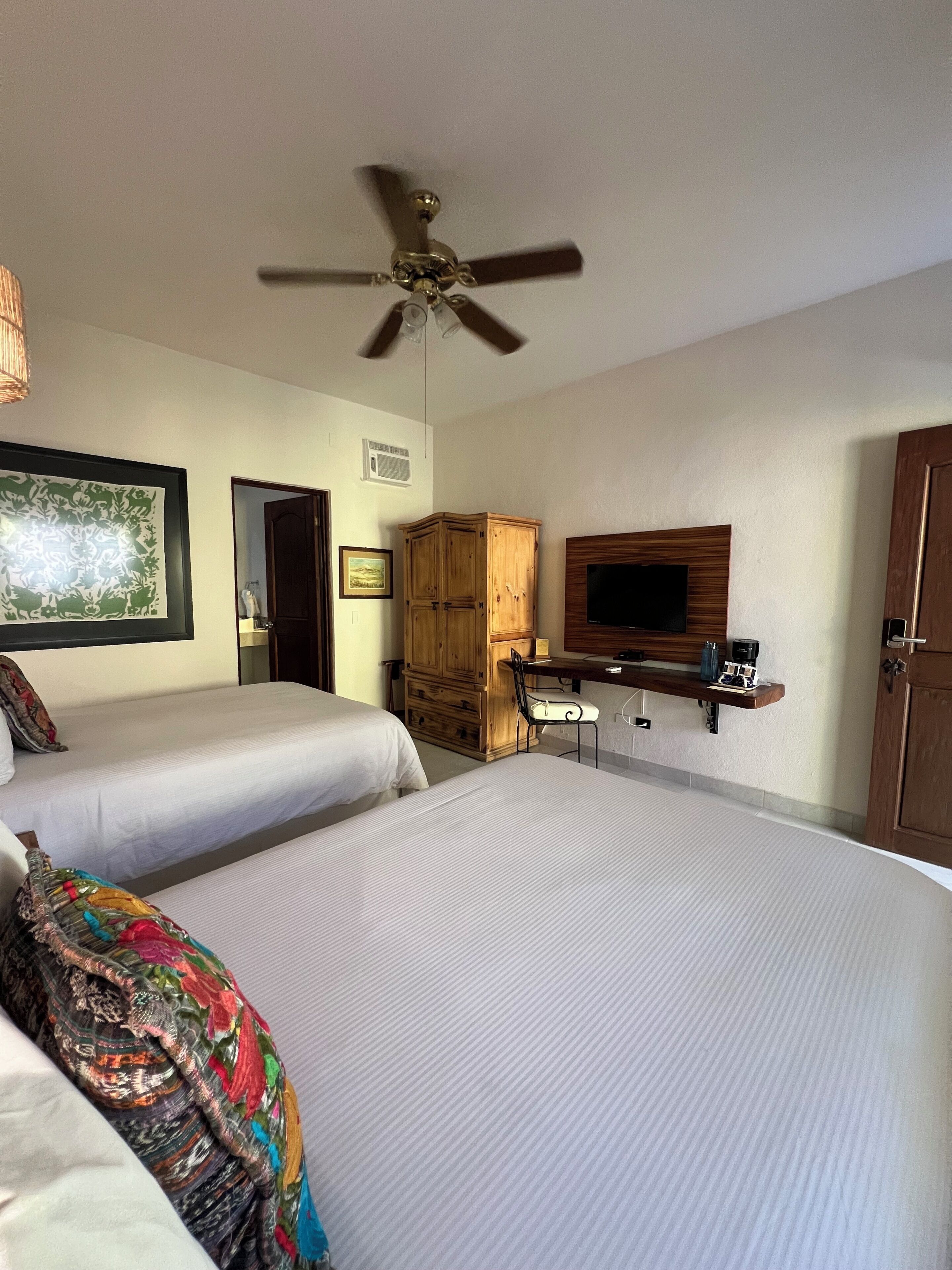 Habitación de hotel con dos camas, un ventilador de techo, un televisor, un armario de madera y un escritorio con una silla.
