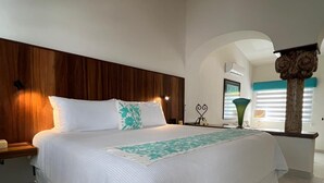 A bedroom with a wooden headboard, a bed with white bedding, a bedside table, a nightstand, a decorative vase, and a window with blinds. - El Encanto Inn (San José del Cabo)