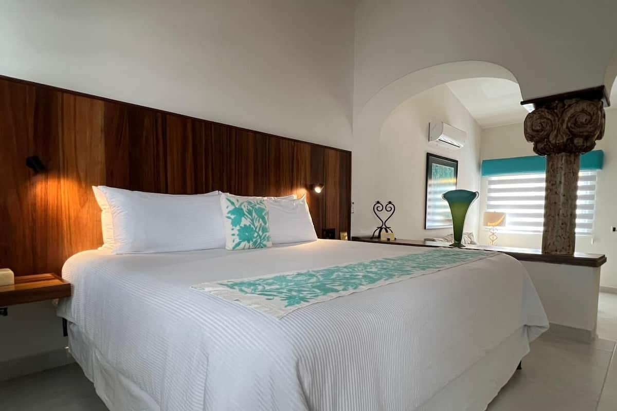 Une chambre à coucher avec une tête de lit en bois, un lit avec une literie blanche, une table de chevet, une commode, un vase décoratif et une fenêtre avec des stores.