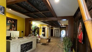 A reception area with a wooden reception desk, a sofa, a decorative wall with a painting, and a plant arrangement. - El Encanto Inn (San José del Cabo)